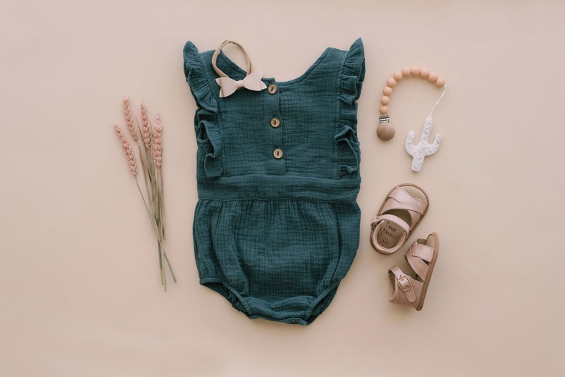 May include: A teal baby onesie with ruffled sleeves and button closures. The onesie is laid flat on a beige background with a wooden teether, sandals, and dried flowers.