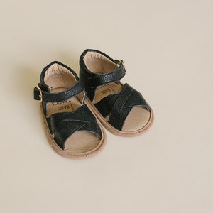 May include: A pair of black leather sandals with a criss-cross strap design. The sandals have a buckle closure and a brown sole. The brand name "BARE BASICS" is printed on the insole.