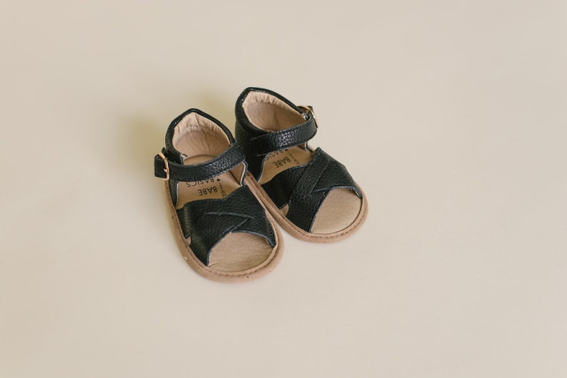 May include: A pair of black leather sandals with a criss-cross strap design. The sandals have a buckle closure and a brown sole. The brand name "BARE BASICS" is printed on the insole.