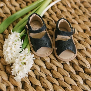 May include: A pair of black leather baby sandals with buckle fastenings. The sandals are on a woven straw surface with white hyacinth flowers.