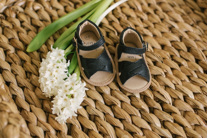 May include: A pair of black leather baby sandals with buckle fastenings. The sandals are on a woven straw surface with white hyacinth flowers.