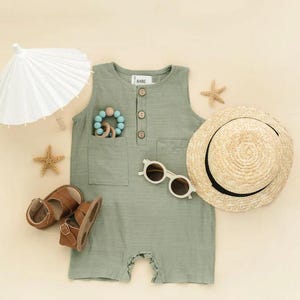 May include: A green sleeveless romper with a button closure and a pocket on the front. The romper is laid out on a beige surface with a straw hat, sunglasses, sandals, a wooden toy, and a white paper parasol.