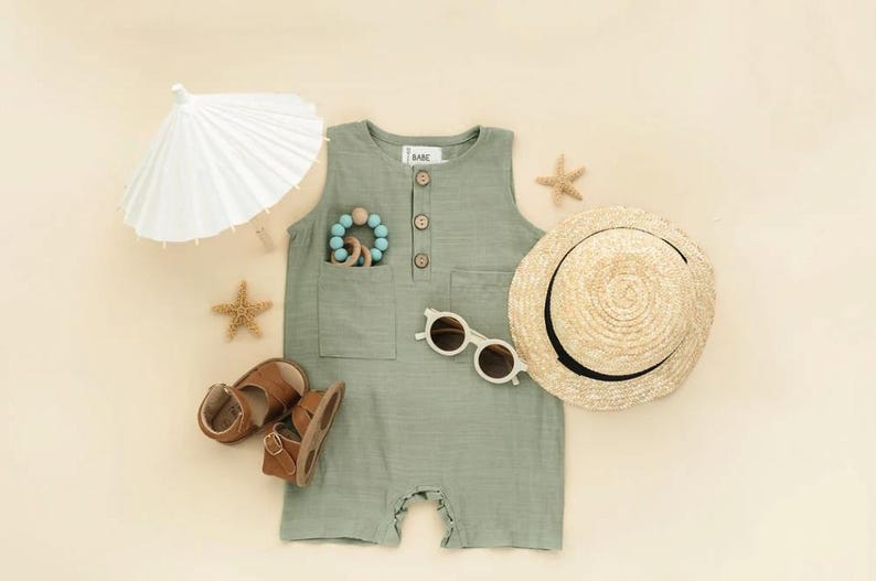 May include: A green sleeveless romper with a button closure and a pocket on the front. The romper is laid out on a beige surface with a straw hat, sunglasses, sandals, a wooden toy, and a white paper parasol.