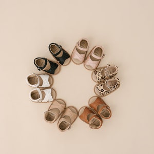 May include: A circle of eight pairs of baby sandals in various colours and patterns, including black, white, brown, pink, and leopard print.