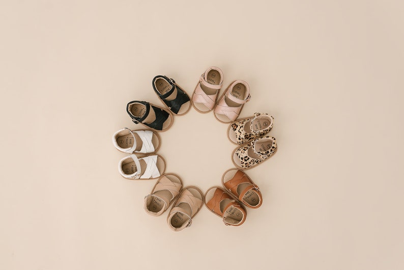 May include: A circle of eight pairs of baby sandals in various colours and patterns, including black, white, brown, pink, and leopard print.