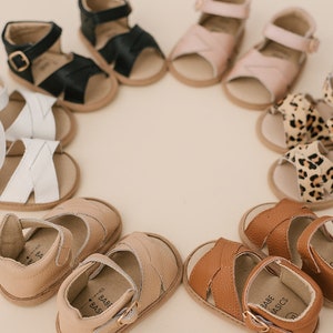 May include: A circle of nine pairs of baby sandals in various colours and patterns, including black, white, tan, brown, pink, and leopard print. The sandals have a buckle fastening and a strap across the toe.