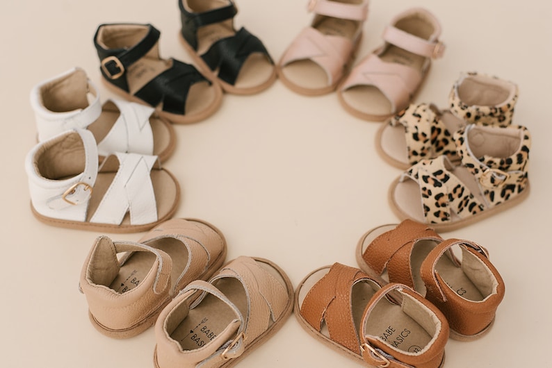 May include: A circle of nine pairs of baby sandals in various colours and patterns, including black, white, tan, brown, pink, and leopard print. The sandals have a buckle fastening and a strap across the toe.