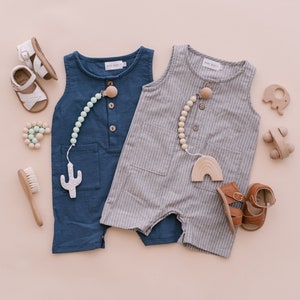 May include: Two baby outfits, one blue and one gray with white stripes, with wooden teethers and sandals. The outfits are sleeveless and have a button closure. The teethers are attached to a string of wooden beads.