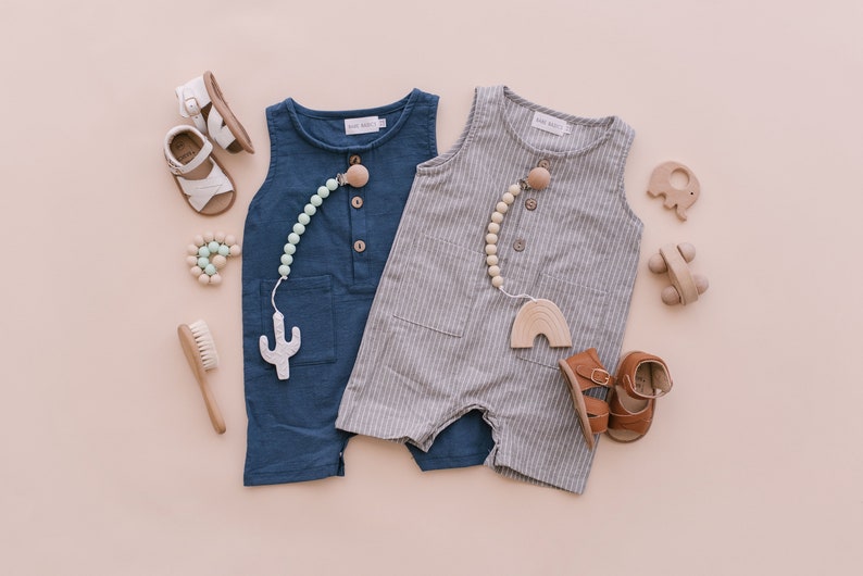 May include: Two baby outfits, one blue and one gray with white stripes, with wooden teethers and sandals. The outfits are sleeveless and have a button closure. The teethers are attached to a string of wooden beads.