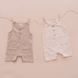 May include: Two sleeveless linen jumpsuits, one beige and one white, hanging on a clothesline against a light pink background.