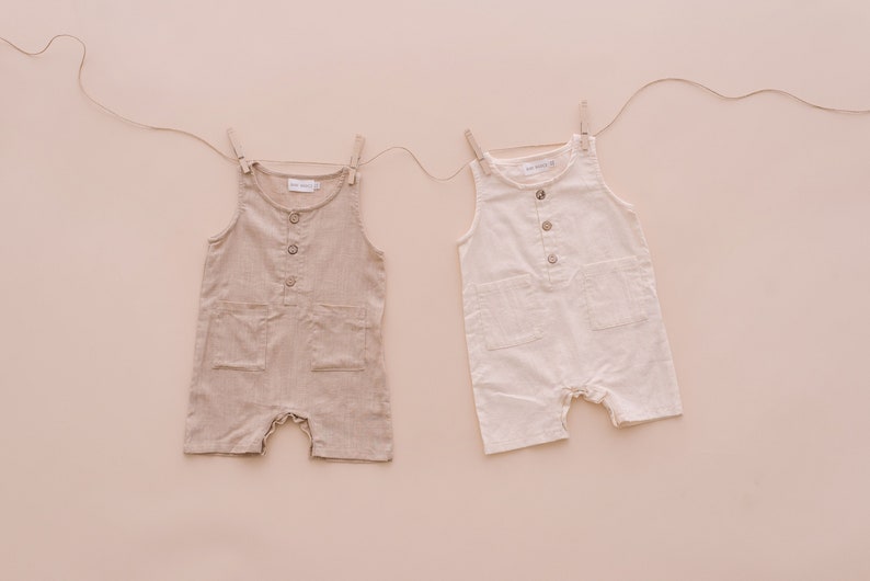 May include: Two sleeveless linen jumpsuits, one beige and one white, hanging on a clothesline against a light pink background.