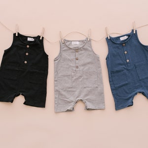 May include: Three sleeveless baby onesies hanging on a clothesline. The onesies are black, gray with white stripes, and blue. Each onesie has a button closure and two pockets.