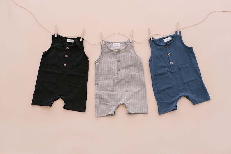 May include: Three sleeveless baby onesies hanging on a clothesline. The onesies are black, gray with white stripes, and blue. Each onesie has a button closure and two pockets.