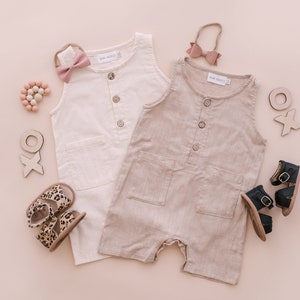 May include: Two sleeveless linen jumpsuits, one white and one beige, with button closures and pockets. The jumpsuits are laid out on a pink background with wooden toys, sandals, and hair accessories.