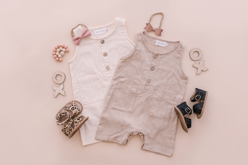 May include: Two sleeveless linen jumpsuits, one white and one beige, with button closures and pockets. The jumpsuits are laid out on a pink background with wooden toys, sandals, and hair accessories.