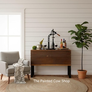 May include: A rustic wooden wine bar with a black metal wine opener, wine bottles, and glasses. A bottle of wine chills in a silver ice bucket. The Painted Cow Shop text is at the bottom. A gray armchair and a round jute rug are in the foreground.