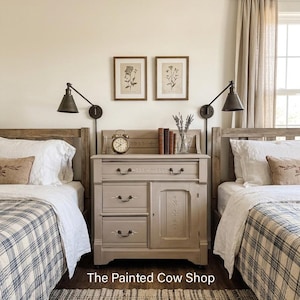 May include: A bedroom scene featuring two wooden beds with plaid bedding. A gray cabinet with drawers and a door sits between the beds, adorned with a clock, books, and a vase. Two framed botanical prints hang above the cabinet. The text "The Painted Cow Shop" is visible.