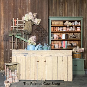 May include: A rustic garden scene featuring a wooden potting bench with gardening tools, watering cans, and a green cabinet filled with terracotta pots. Hydrangeas in various containers add a touch of color. The Painted Cow Shop text is visible.