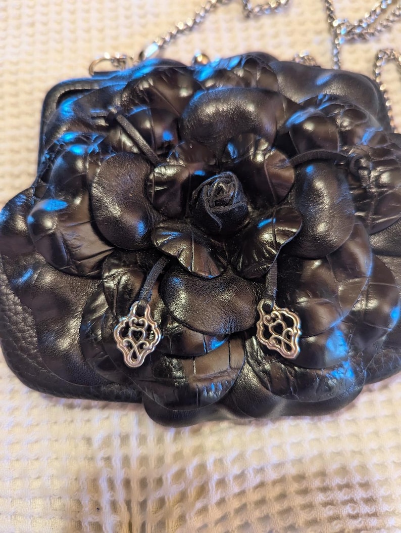 K&ouml;nnte beinhalten: Schwarze Lederhandtasche in Form einer Rose mit silbernen Akzenten. Die Tasche hat einen Kettenriemen.