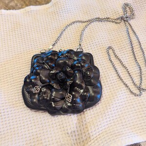 K&ouml;nnte beinhalten: Eine schwarze Ledertasche in Form einer Blume mit einem silbernen Kettenriemen.