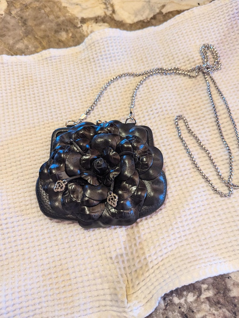 K&ouml;nnte beinhalten: Eine schwarze Ledertasche in Form einer Blume mit einem silbernen Kettenriemen.