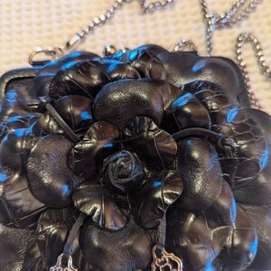 K&ouml;nnte beinhalten: Eine schwarze Lederhandtasche mit einem gro&szlig;en Blumendesign. Die Handtasche hat einen silbernen Kettenriemen und silberne Akzente.