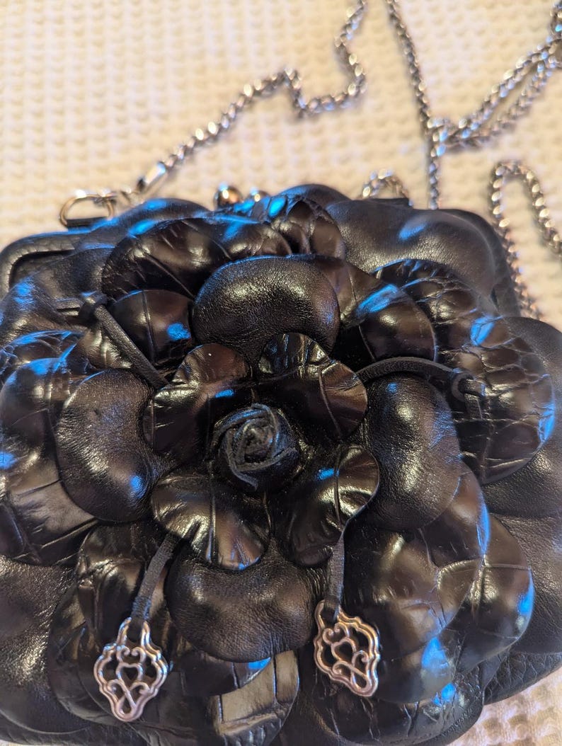 K&ouml;nnte beinhalten: Eine schwarze Lederhandtasche mit einem gro&szlig;en Blumendesign. Die Handtasche hat einen silbernen Kettenriemen und silberne Akzente.