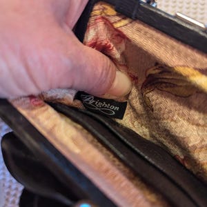 K&ouml;nnte beinhalten: Eine schwarze Leder-Clutch mit floralem Futter und einem silbernen Verschluss. Die Clutch hat ein Brighton-Markenetikett im Inneren.