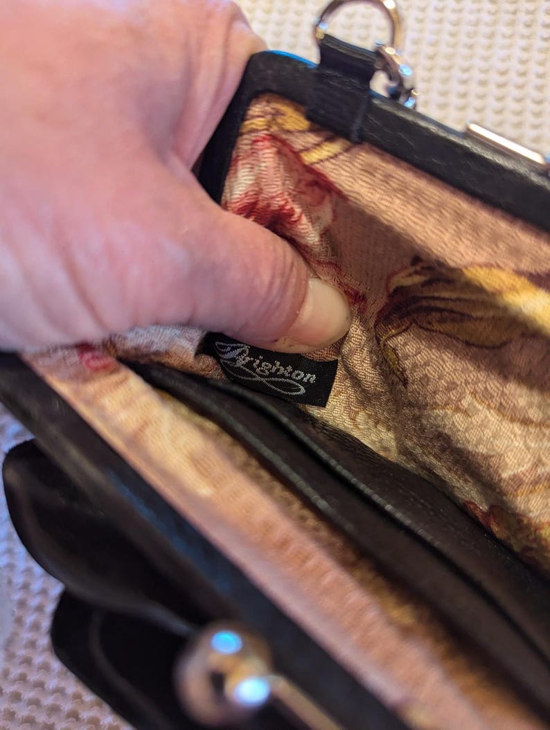 K&ouml;nnte beinhalten: Eine schwarze Leder-Clutch mit floralem Futter und einem silbernen Verschluss. Die Clutch hat ein Brighton-Markenetikett im Inneren.