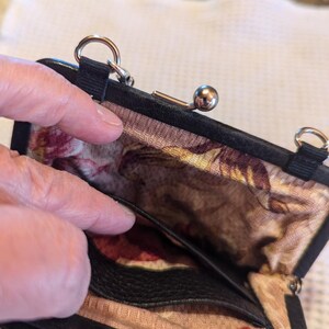 K&ouml;nnte beinhalten: Eine schwarze Lederm&uuml;nzb&ouml;rse mit einem floralen Stofffutter und einem silbernen Verschluss. Die B&ouml;rse hat eine kleine Tasche im Inneren und einen Kettenriemen.