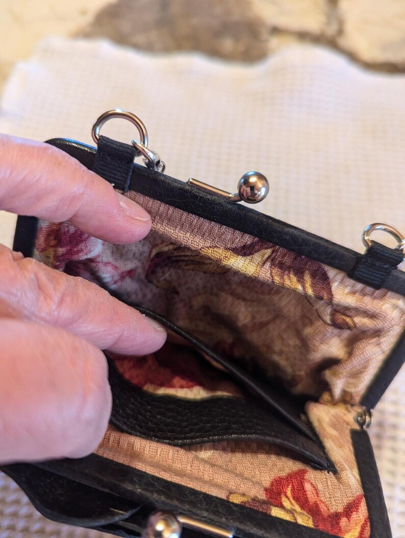 K&ouml;nnte beinhalten: Eine schwarze Lederm&uuml;nzb&ouml;rse mit einem floralen Stofffutter und einem silbernen Verschluss. Die B&ouml;rse hat eine kleine Tasche im Inneren und einen Kettenriemen.
