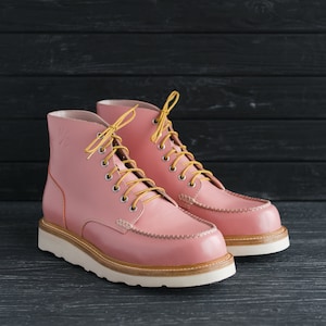 May include: A pair of pink leather boots with yellow laces. The boots have a white sole and a moc toe design.