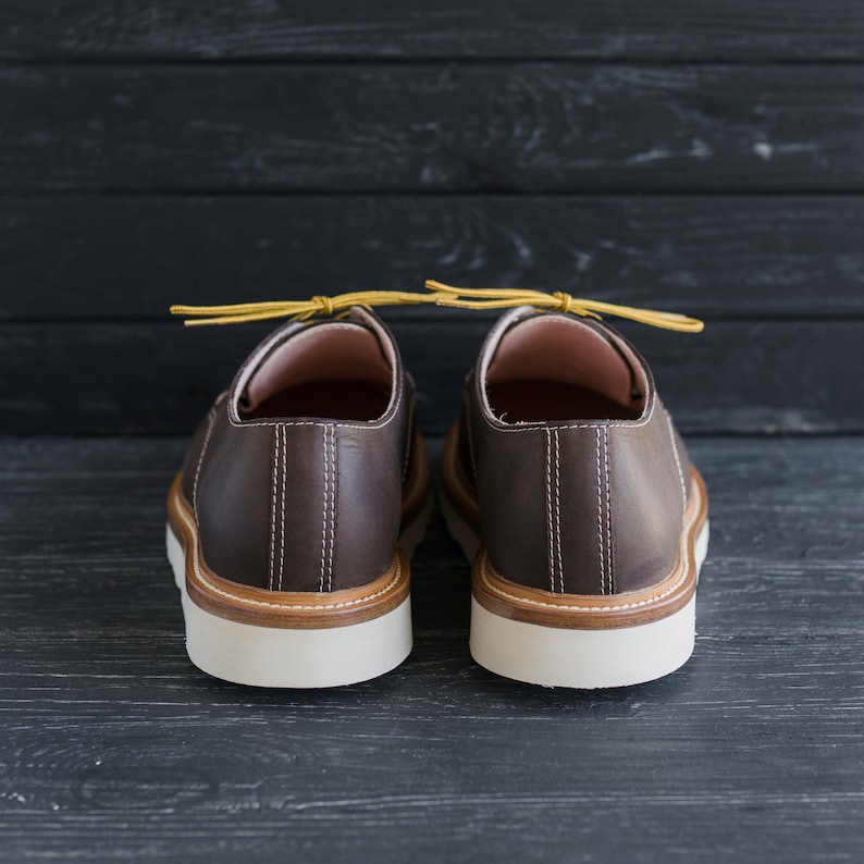 May include: A pair of brown leather shoes with yellow laces. The shoes have a white sole and are on a black wooden background.