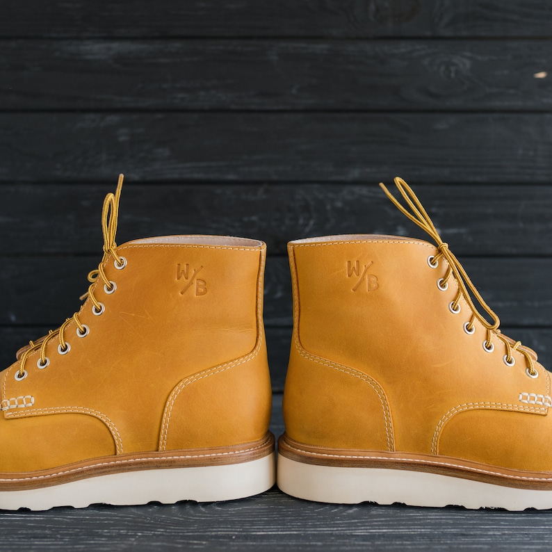 Puede incluir: Un par de botas de cuero amarillo con suelas y cordones blancos. Las botas tienen un logotipo en el lateral que dice "W/B".