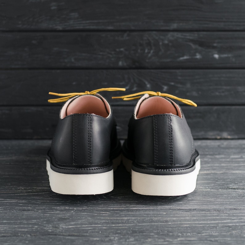 Peut inclure: Une paire de chaussures en cuir noir avec des semelles blanches et des lacets jaunes. Les chaussures sont vues de l'arri&egrave;re.