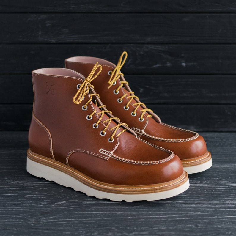 May include: A pair of brown leather boots with yellow laces. The boots have a white rubber sole and a moc toe design.