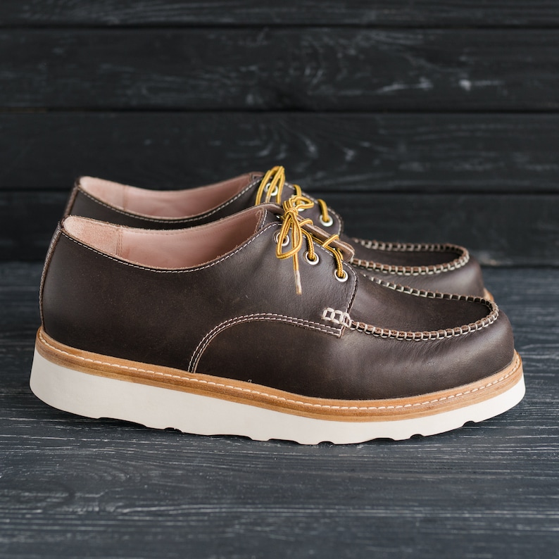 May include: A pair of brown leather lace-up shoes with a white rubber sole. The shoes have a moc toe design and yellow stitching detail.