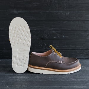 May include: Brown leather lace-up shoe with a white sole and a yellow lace. The shoe is on a black wooden surface. The sole of the shoe is visible and has the text "Vibram" and "Treadlex" on it.
