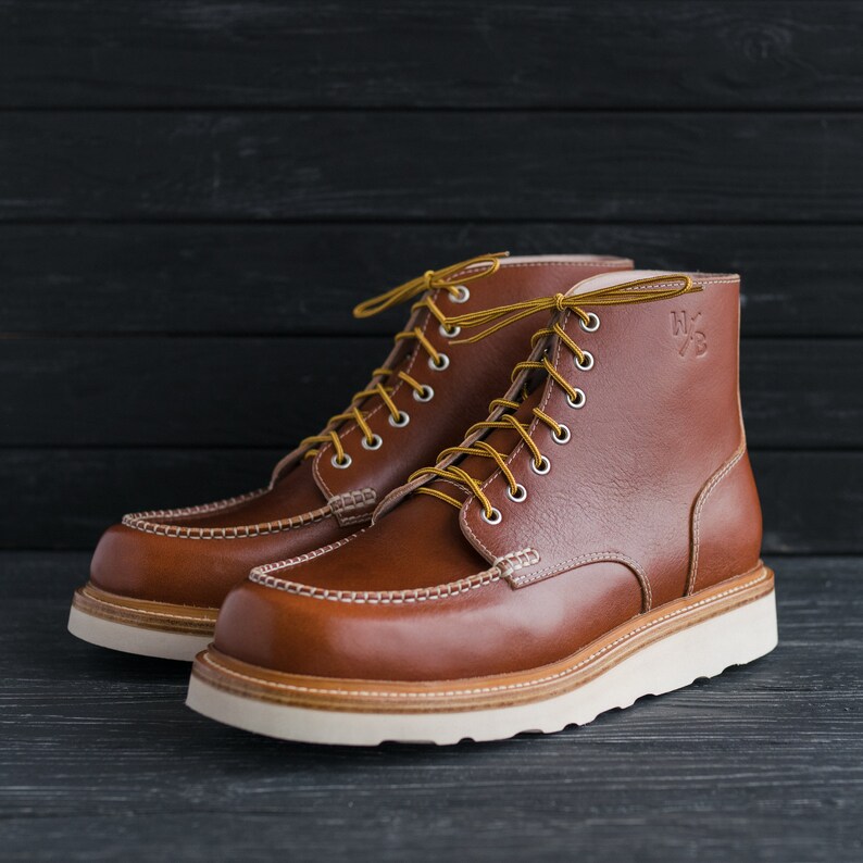 May include: A pair of brown leather boots with white soles and yellow laces. The boots have a moc toe design and a  "W.L.  " logo on the side.