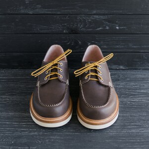 May include: A pair of brown leather boat shoes with yellow laces. The shoes have a rounded toe and a white rubber sole.