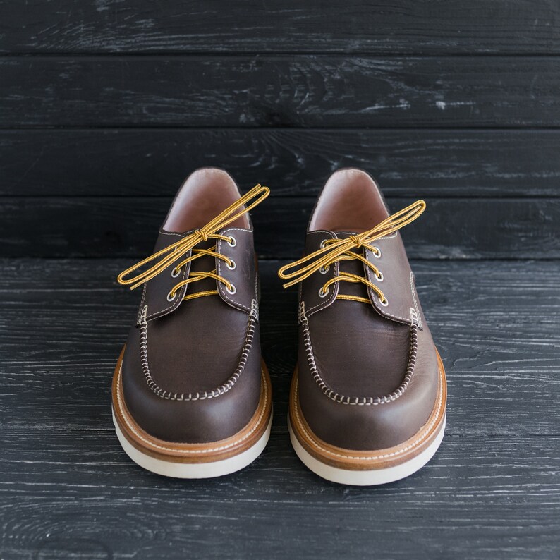 May include: A pair of brown leather boat shoes with yellow laces. The shoes have a rounded toe and a white rubber sole.