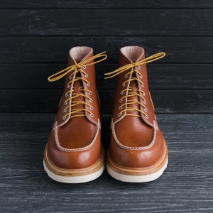 May include: A pair of brown leather boots with yellow laces. The boots have a white sole and are on a black wooden background.