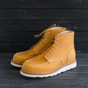 Puede incluir: Un par de botas de trabajo de cuero marr&oacute;n con cordones blancos y suela blanca. Las botas tienen una costura cosida alrededor de la punta y un peque&ntilde;o logotipo en el lateral.