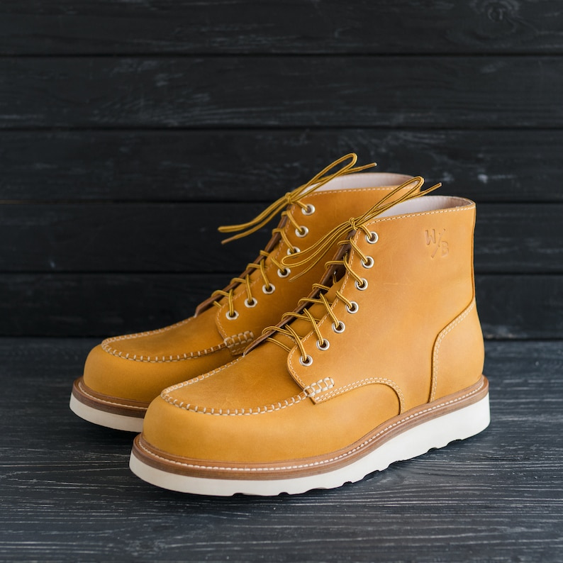 Puede incluir: Un par de botas de trabajo de cuero marr&oacute;n con cordones blancos y suela blanca. Las botas tienen una costura cosida alrededor de la punta y un peque&ntilde;o logotipo en el lateral.