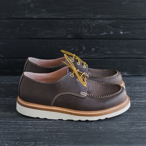 May include: A pair of brown leather lace-up shoes with a white rubber sole. The shoes have a moc toe design and are tied with yellow laces.