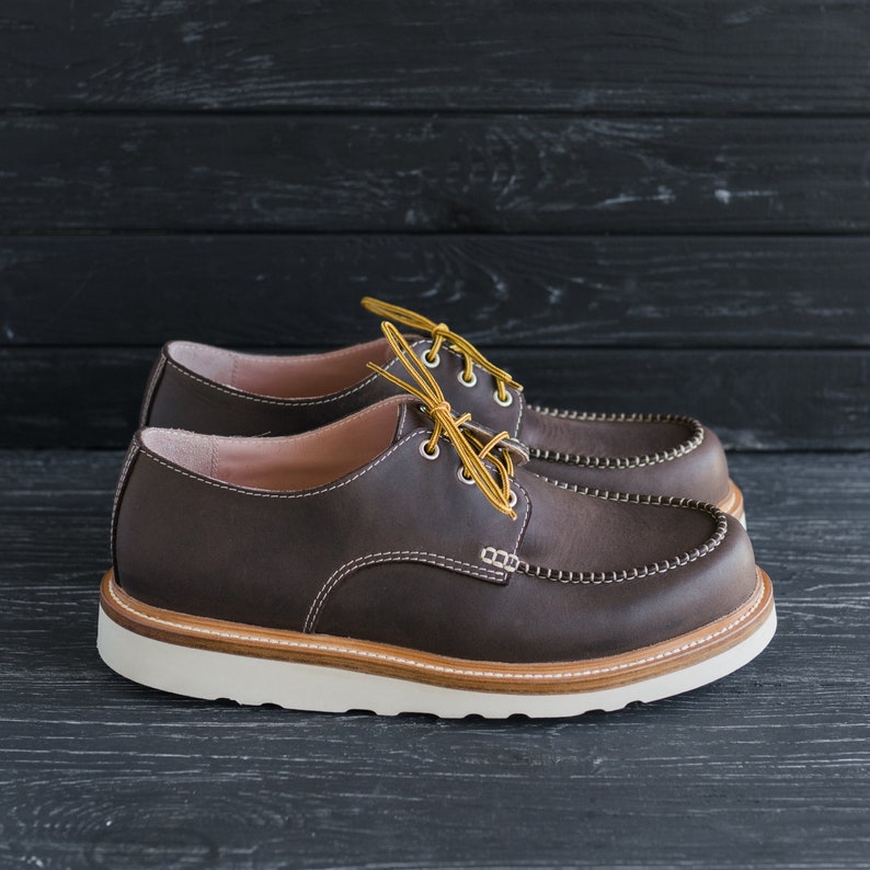 May include: A pair of brown leather lace-up shoes with a white rubber sole. The shoes have a moc toe design and are tied with yellow laces.