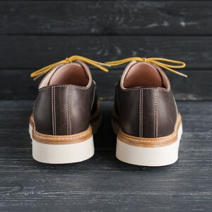 May include: A pair of brown leather lace-up shoes with yellow laces. The shoes have a white sole and are on a dark wooden background.