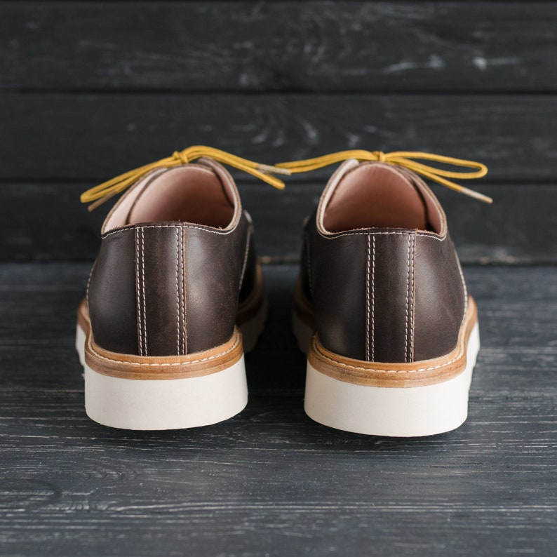May include: A pair of brown leather lace-up shoes with yellow laces. The shoes have a white sole and are on a dark wooden background.