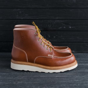 May include: Brown leather boots with white soles and laces. The boots have a stitched seam around the toe and a logo on the side that says "W.E."