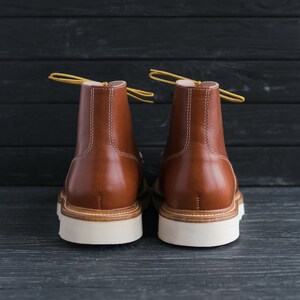 May include: A pair of brown leather boots with white soles. The boots have yellow laces and are shown from the back.